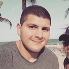 A person smiles at the camera with a beach background, clear skies, and palm trees. They have short hair and are wearing a gray t-shirt.