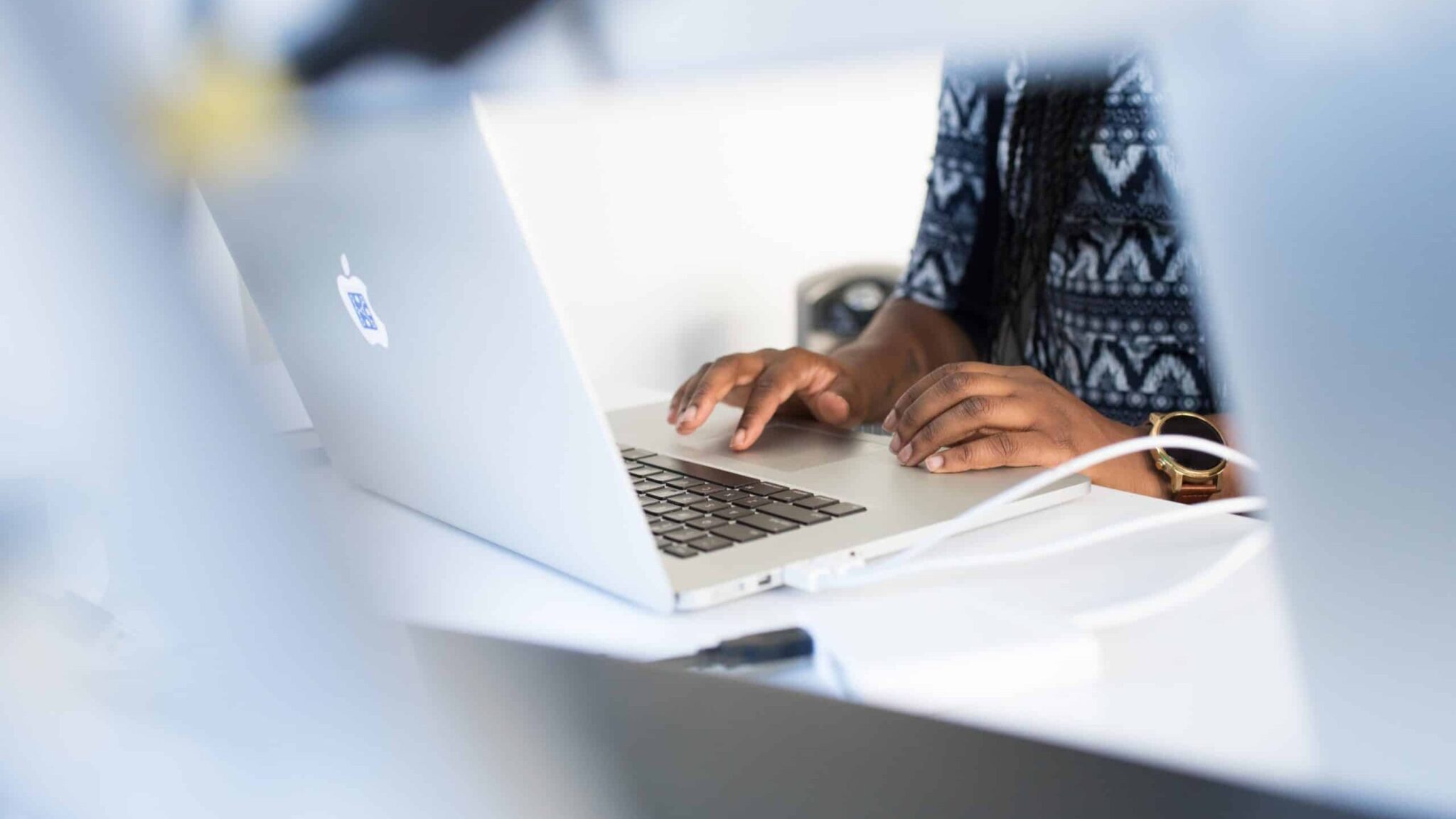 A person is working on a laptop with white earphones. Focus is on their hands typing. The environment appears to be a light workspace.