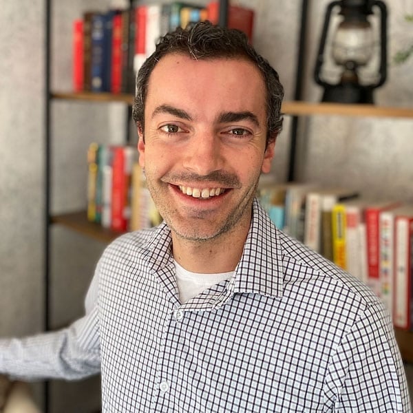 A smiling person wears a checkered shirt and stands in front of a bookshelf filled with colorful books. The setting appears informal and friendly.
