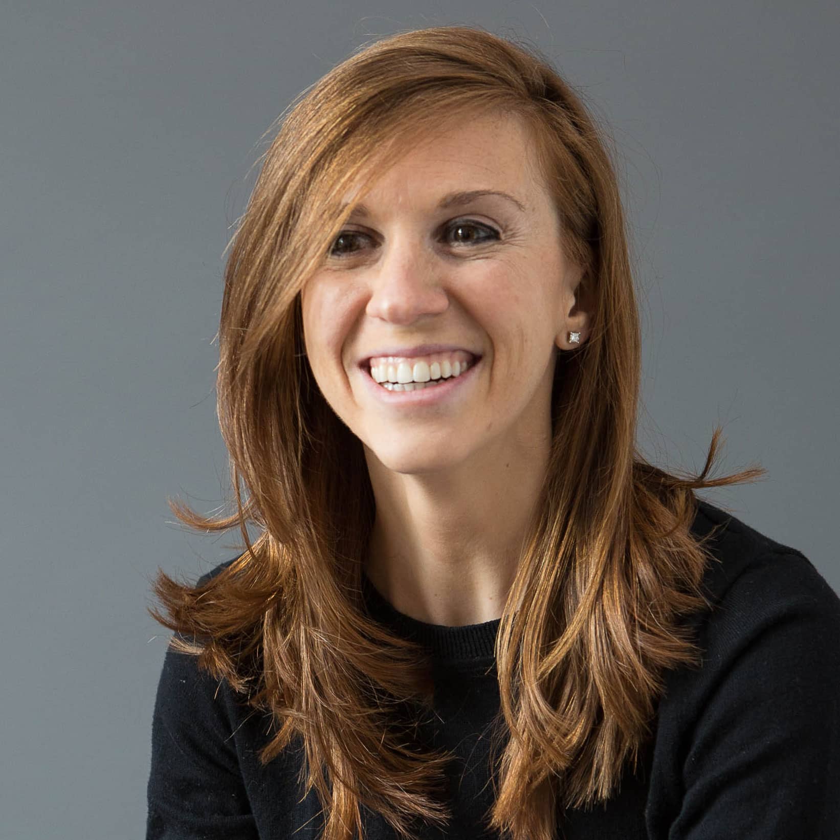 This image shows a person with shoulder-length brown hair smiling at an angle against a gray background, wearing a black top and stud earrings.