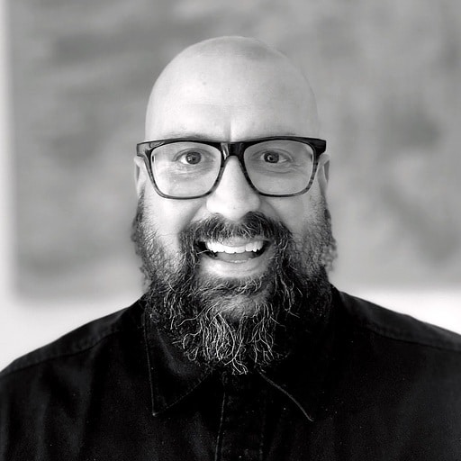 The image is a black and white portrait of a smiling person with a bald head, thick beard, glasses, and a black shirt, with a blurred background.
