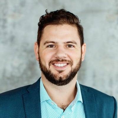 A smiling person with a beard, wearing a blue blazer and a patterned shirt, stands against a textured gray background. Professional headshot style.