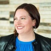 A person with shoulder-length hair smiling, wearing a blue top, black leather jacket, and earrings, with a blurred brick wall background.