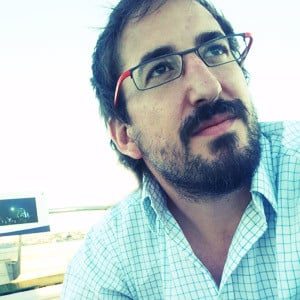The image shows a person with facial hair wearing red-rimmed glasses and a blue checkered shirt, looking upward thoughtfully against a blurred background.