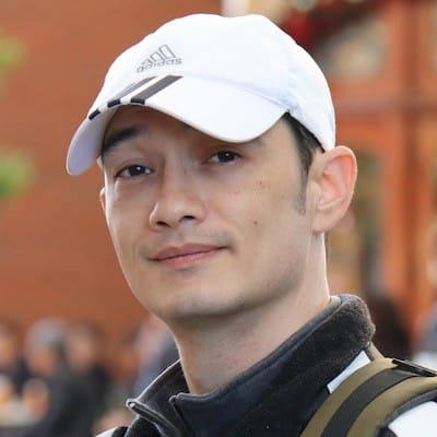 A person is wearing a white Adidas cap, looking at the camera with a slight smile. They have a backpack strap visible and a black jacket.