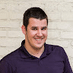 The image shows a smiling person wearing a purple polo shirt, posed against a white brick wall, exuding a friendly and approachable demeanor.