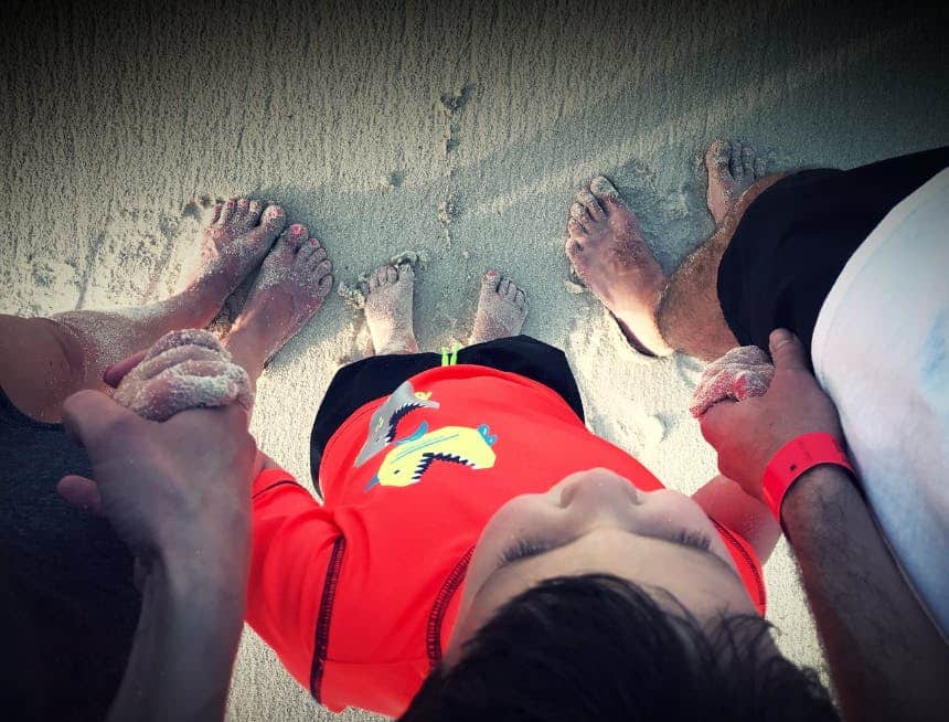 Three people are standing on a sandy beach with their bare feet visible, showing two adults and a child wearing an orange shirt with a dinosaur design.