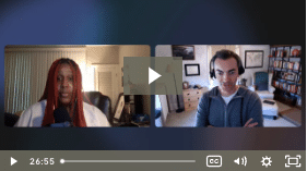 A video conference screenshot with two people, one with red hair, another wearing a grey top. Both are indoors, and it's a paused video with a play button.