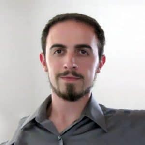 The image shows a person with short dark hair and a beard, wearing a gray shirt, against a mostly white background. The person is looking at the camera.