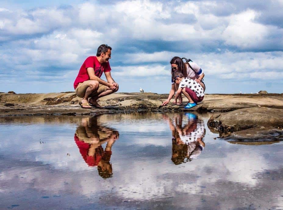 Two people are exploring a shallow water pool on a rocky landscape under a cloudy sky, reflecting on the water's surface.