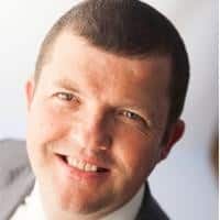 Close-up of a person smiling at the camera, wearing a suit without a tie. The person has short hair, and the background is out of focus.