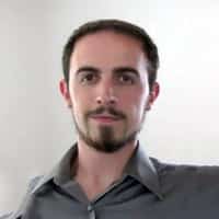 A person with short dark hair and a beard, wearing a gray shirt, is standing against a white background, looking directly at the camera.