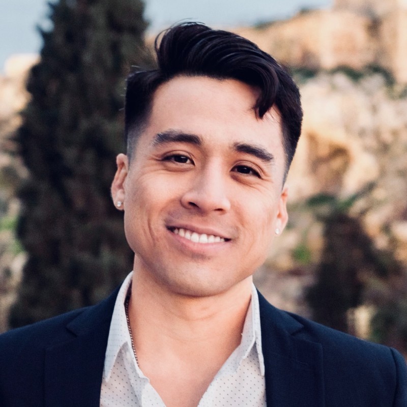 The image shows a smiling person with short hair, wearing a suit without a tie, outdoors with trees and a pale sky in the background.