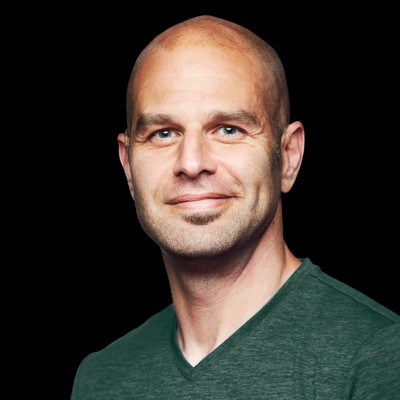 This is a close-up image of a smiling person with a bald head, stubble, and blue eyes wearing a dark green V-neck shirt against a black background.