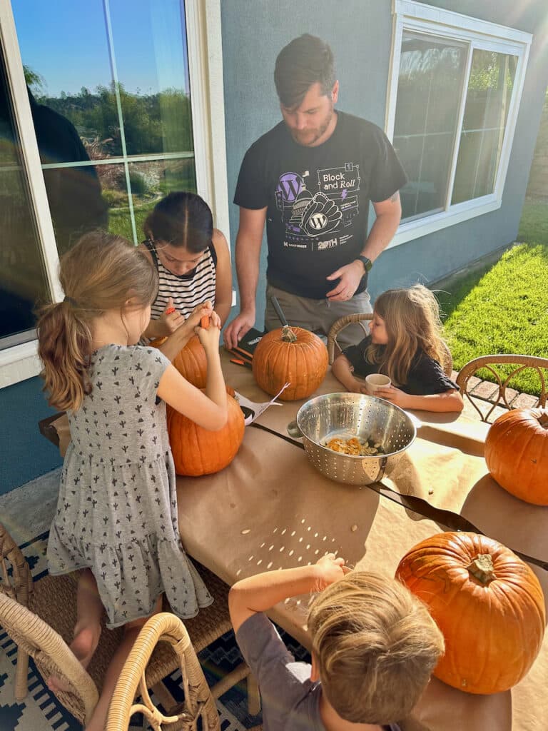 A person and children are carving pumpkins on a sunny day, with a colander of pumpkin seeds and tools on the table.