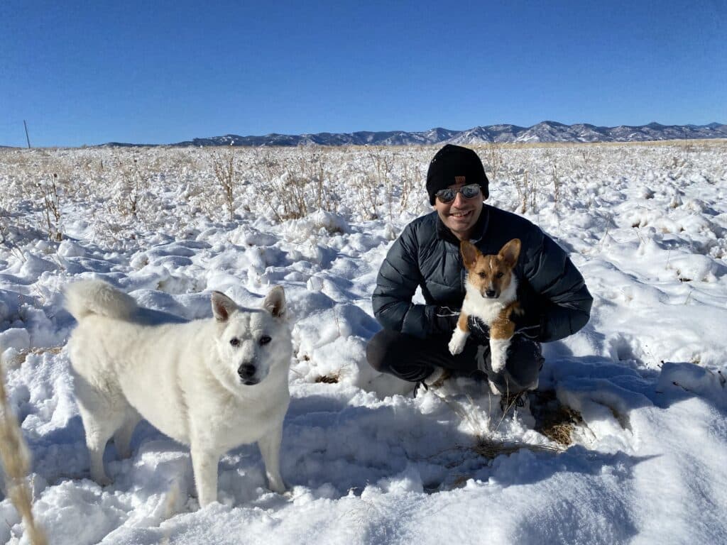 A person is crouching in the snow with two dogs, one white and one brown, against a backdrop of mountains under a clear blue sky.