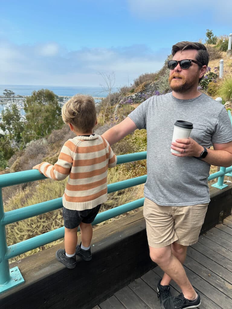 A person and a child stand by a railing overlooking a coastal landscape, with the adult holding a coffee cup, both dressed casually.