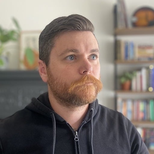 A person with a styled beard is gazing to the side. They're wearing a black hoodie and behind them is a blurred bookshelf.