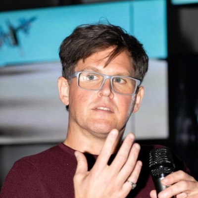 A person with glasses holding a microphone gestures while speaking. A blurred background features a turquoise abstract image, possibly projected.