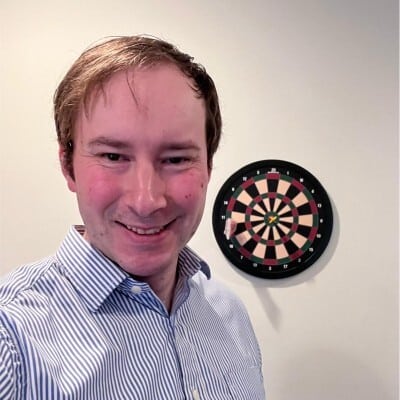 A person in a striped shirt stands indoors, smiling. A dartboard is visible on the wall behind them.
