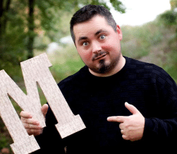 A person in a black sweater holds a large letter "M" in an outdoor setting with trees in the background.