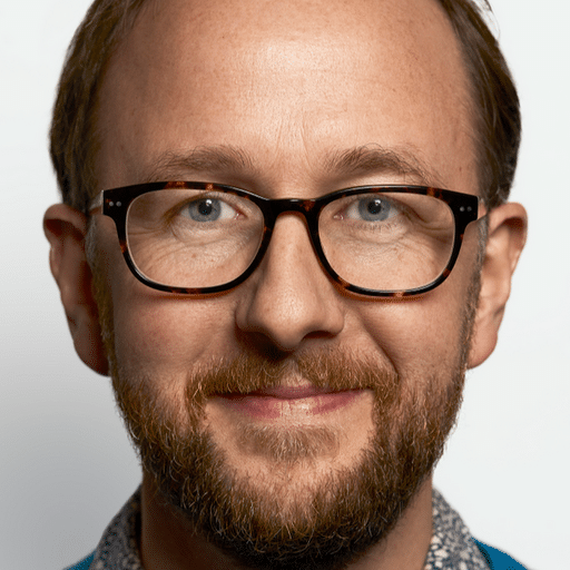 A person wearing glasses, smiling, with a beard and blue jacket.
