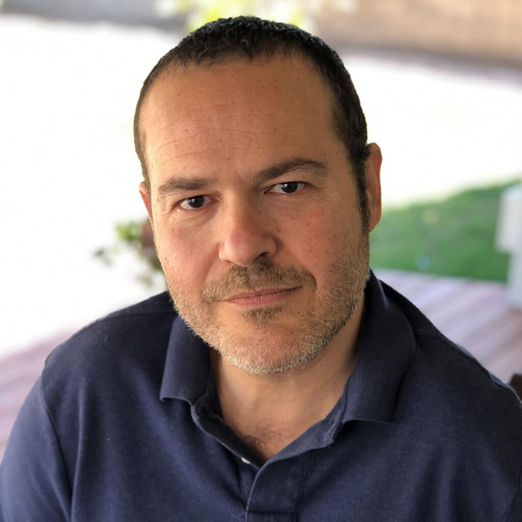 A person with short hair and facial hair is outdoors, wearing a dark-collared shirt, looking into the camera. Blurred greenery in background.