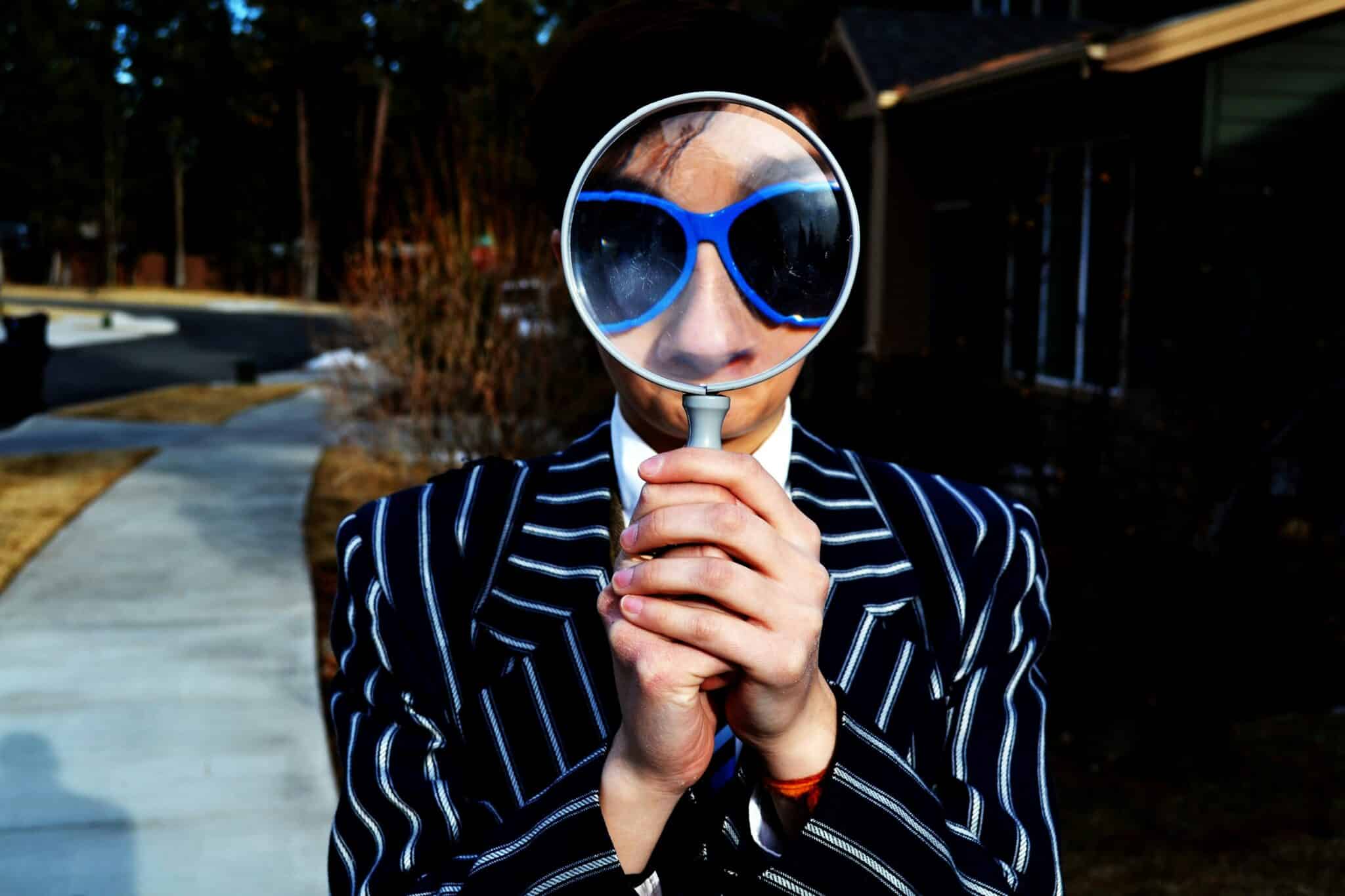A person in a striped suit holds a magnifying glass over sunglasses, distorting facial features. Trees and a path are visible in the background.