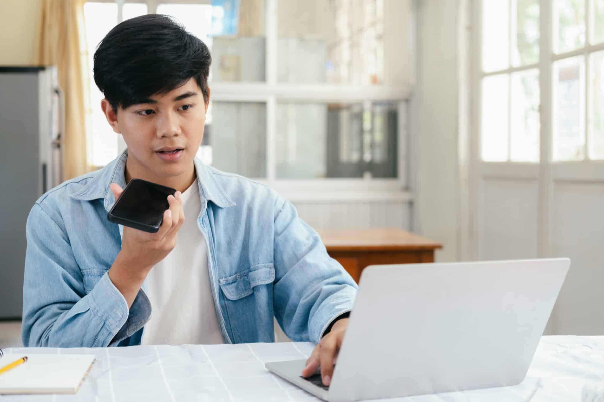 A person in a denim shirt uses a smartphone and laptop at a table, appearing focused in a bright, minimalist room.