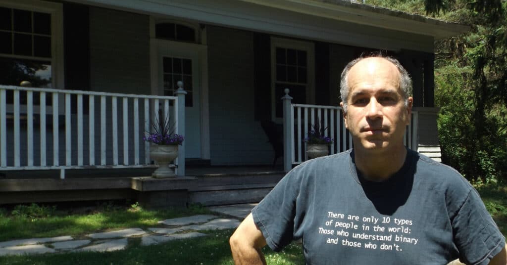 Person standing in front of a porch with potted plants, wearing a shirt with a binary code joke. Wooden steps lead to the entrance.