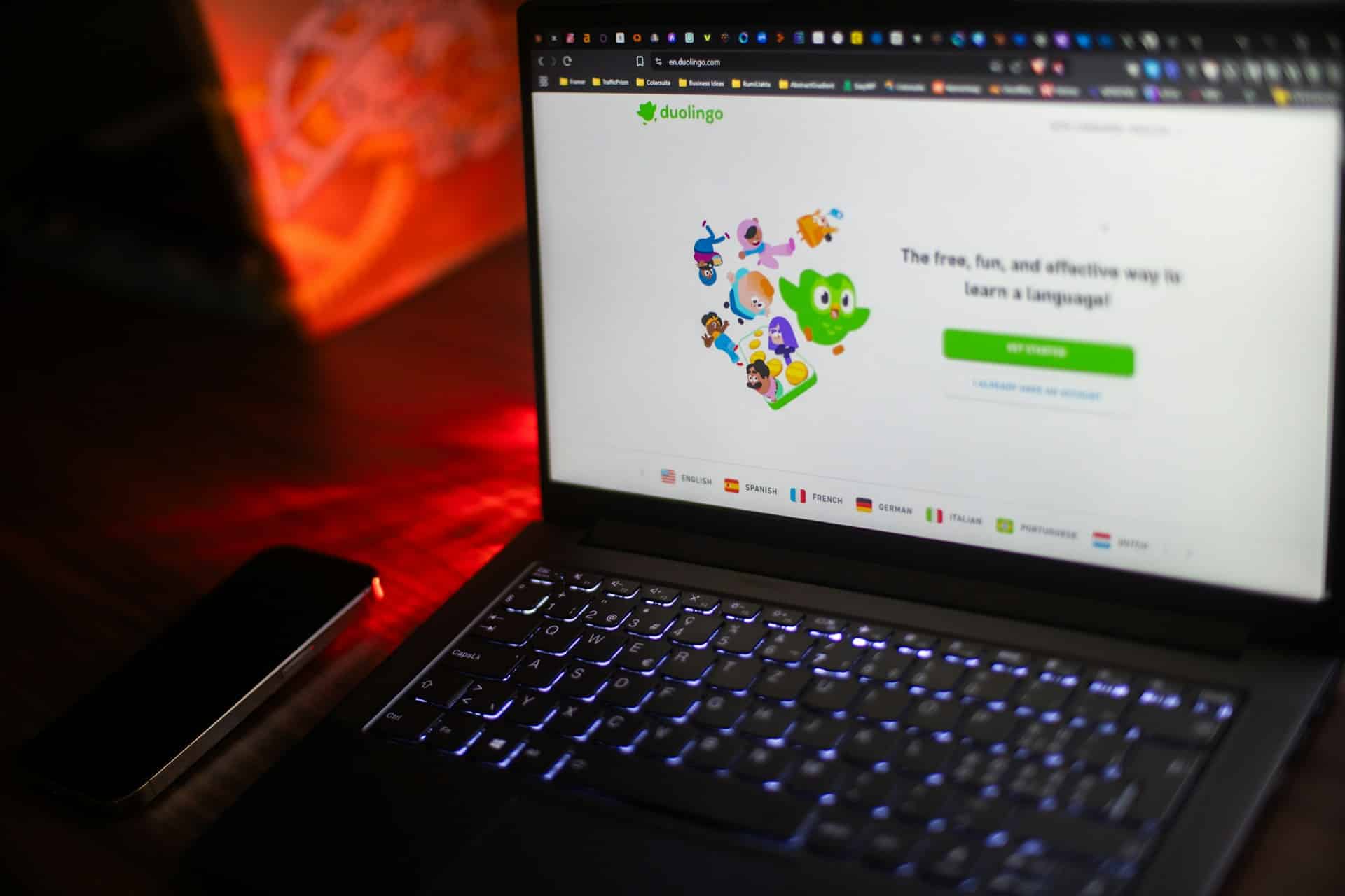 A laptop displays the Duolingo website, surrounded by a dark ambiance with a smartphone nearby, creating a focused study setup.