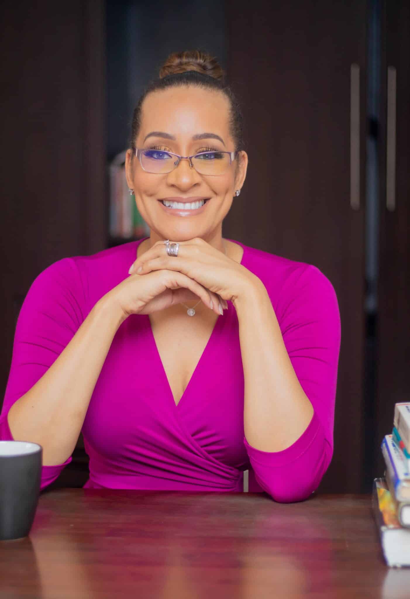 A person wearing glasses and a bright pink top smiles in a cozy room, seated at a table with a cup and books.