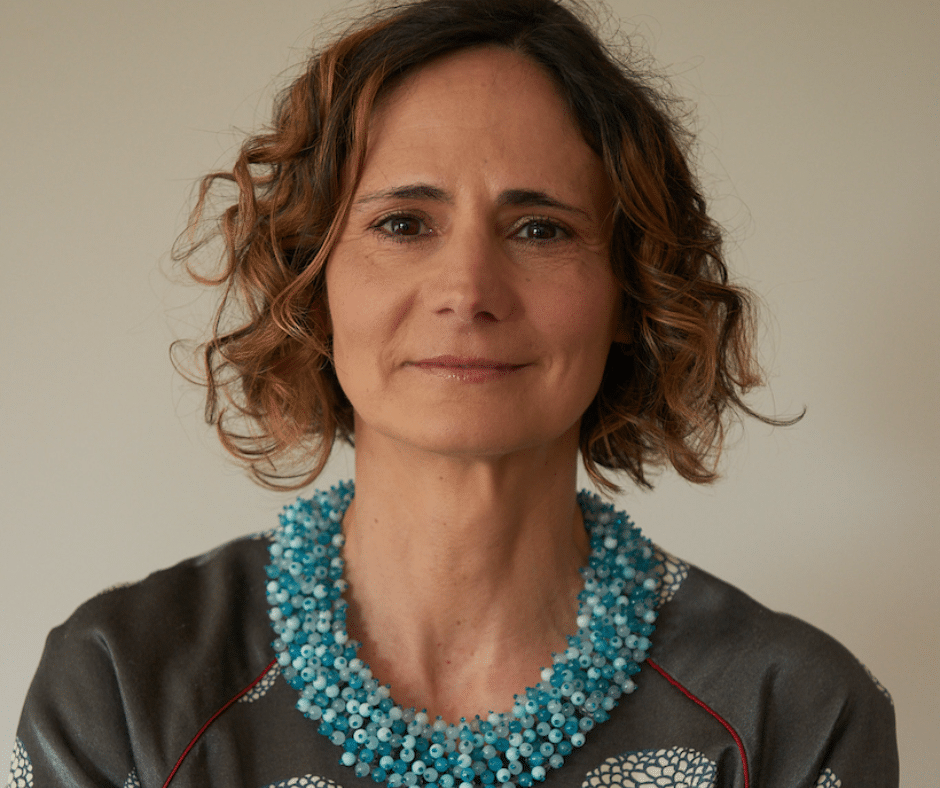 A person with curly hair wears a blue necklace and patterned top, smiling slightly against a plain background.
