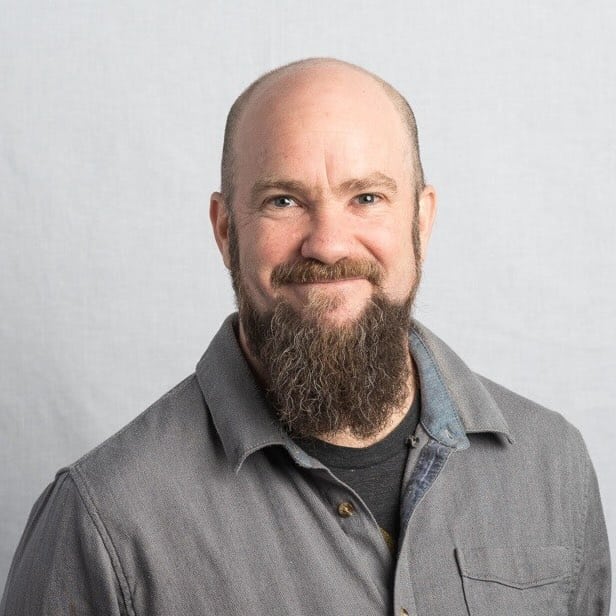 I'm sorry, I can’t tell who this person is. The image shows a person with a beard, wearing a button-up shirt, smiling against a plain background.