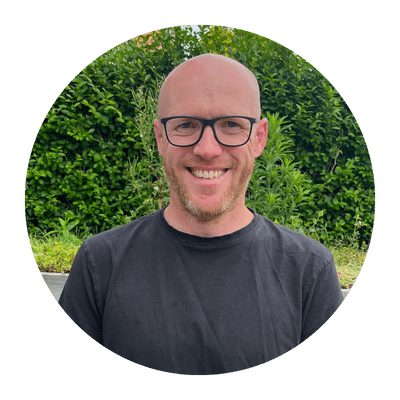A person smiling, wearing glasses and a black shirt, stands outdoors in front of greenery.