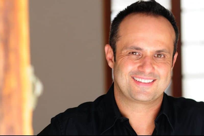 A person smiles brightly, wearing a black shirt, indoors with a blurred background featuring a wooden door and window blinds.