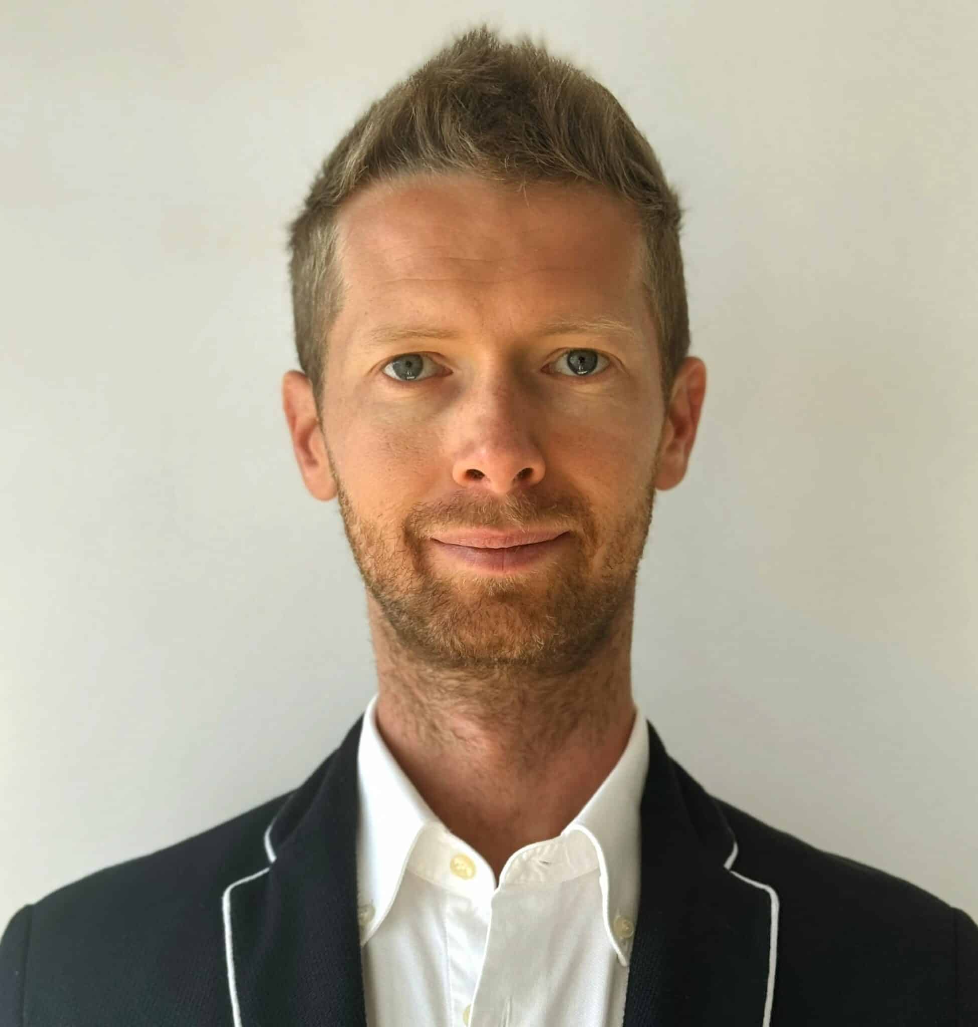 A person with short hair and a beard wears a suit with a white shirt, standing against a plain white background.
