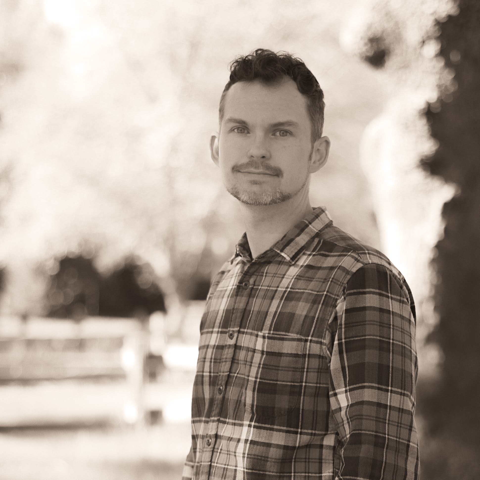 A person in a plaid shirt stands outdoors against a blurred background, featuring trees and wooden fencing, in a sepia-toned photograph.