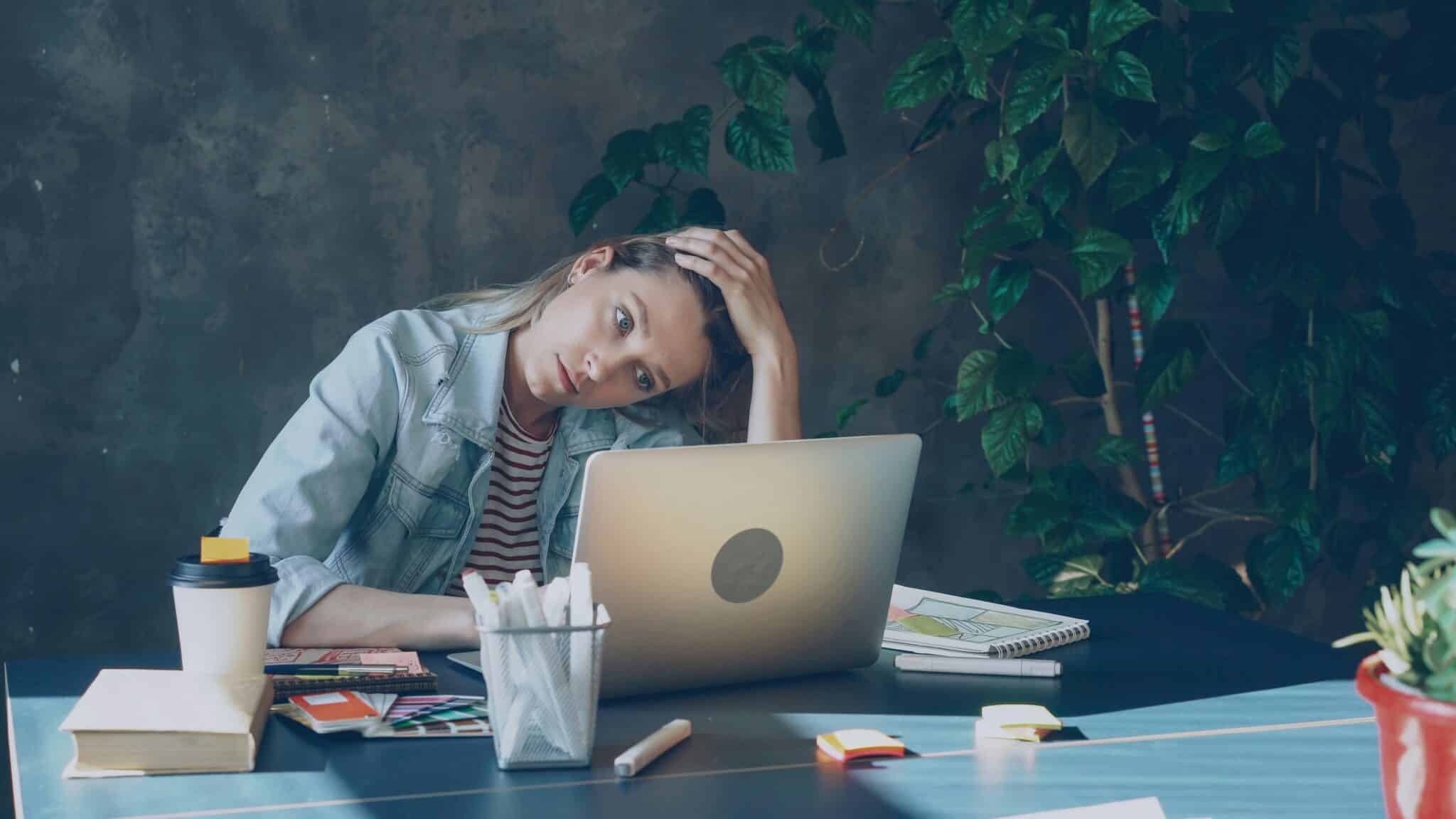 A person sits at a desk with a laptop, surrounded by plants, books, stationery, and a coffee cup, looking contemplative.