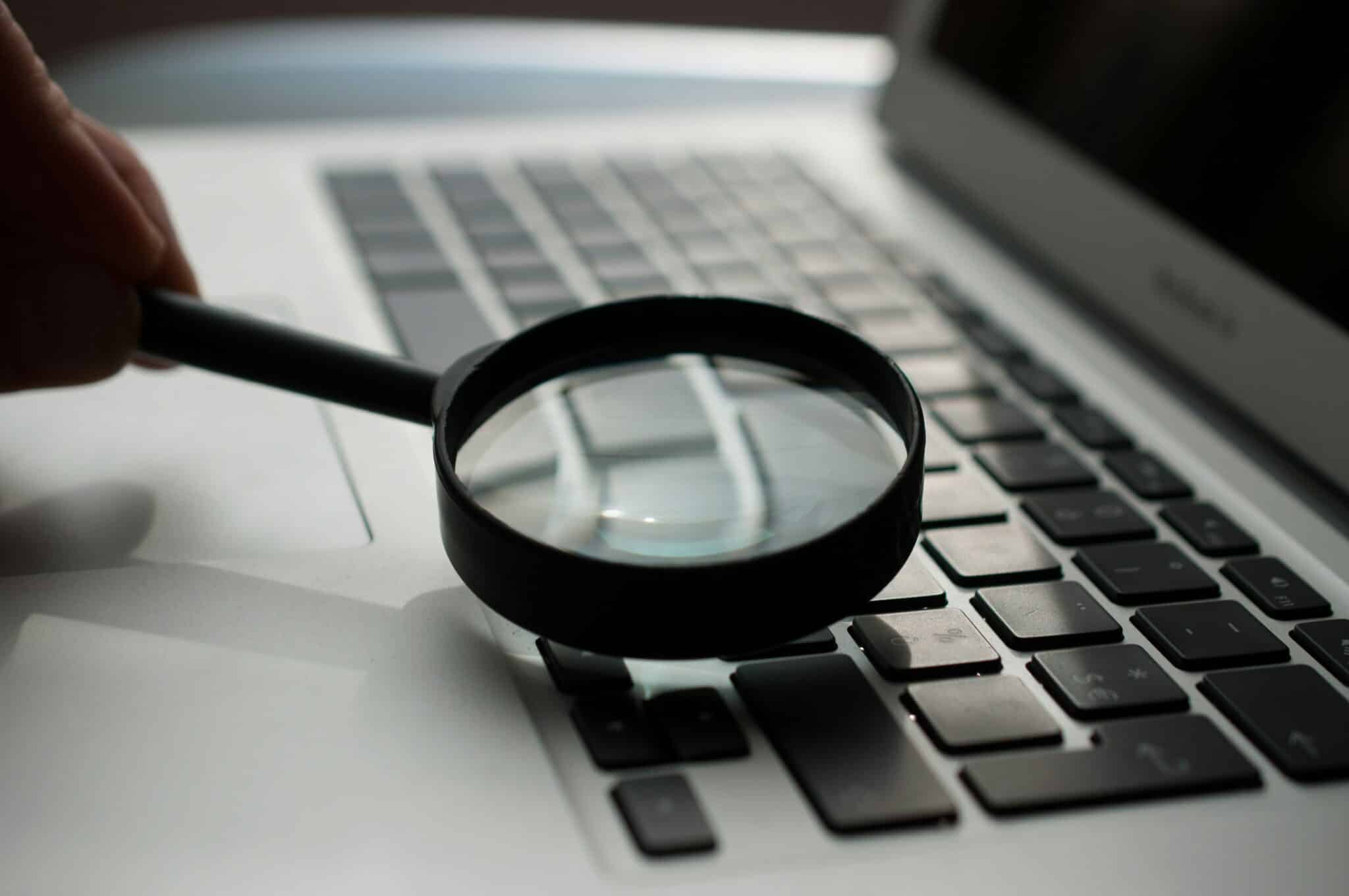 A magnifying glass is held over a laptop keyboard, focusing on the keys, suggesting a search or detailed inspection concept.