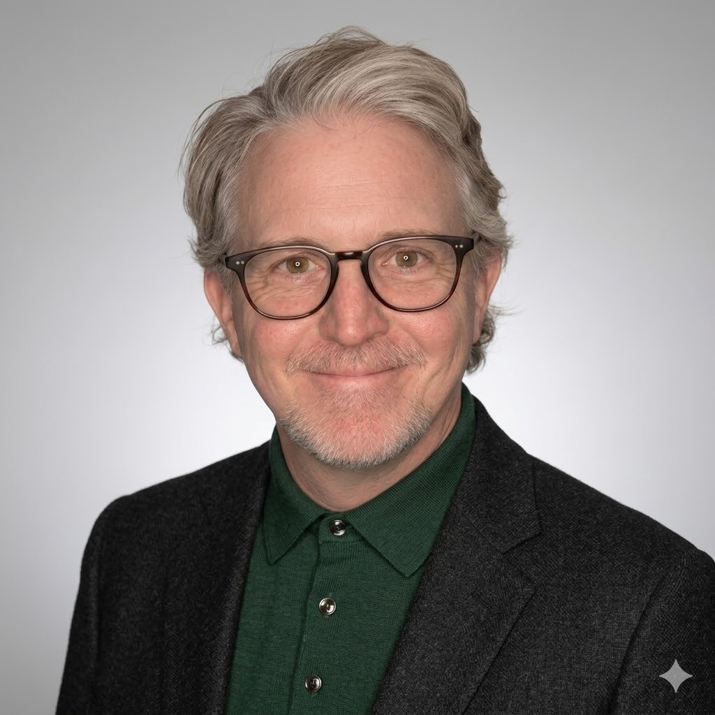 Portrait of a smiling middle-aged man with gray hair and glasses, wearing a dark blazer and green shirt against a light gray background.