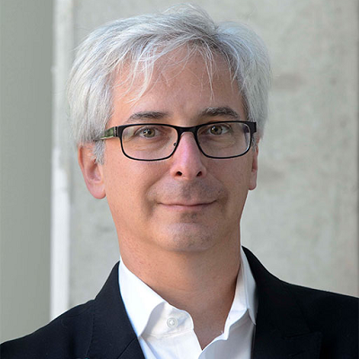 Close-up portrait of a man with gray hair and glasses, wearing a dark suit and white shirt, looking at the camera.