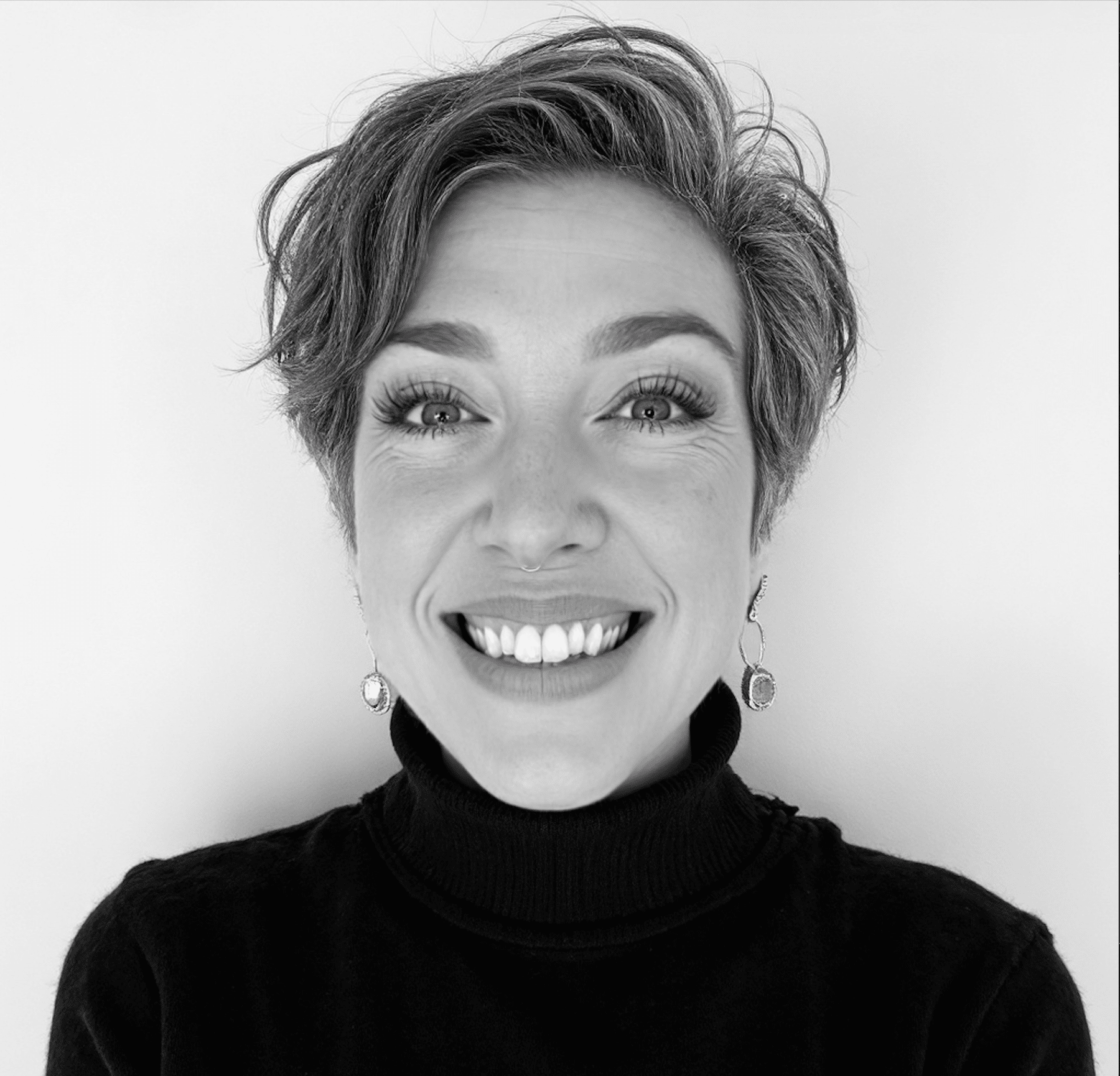 Black-and-white portrait of a smiling woman with short hair, wearing a black turtleneck and dangling earrings.