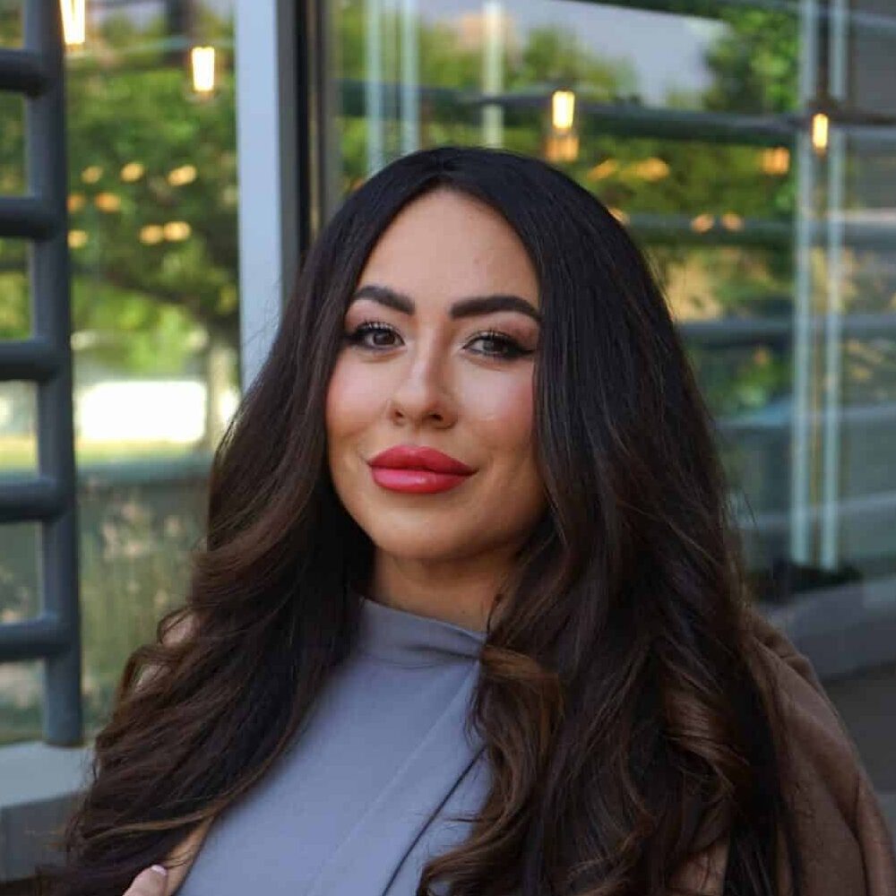 A woman with long dark hair wearing a lavender dress and a brown coat draped over her shoulders, standing on a rooftop patio.