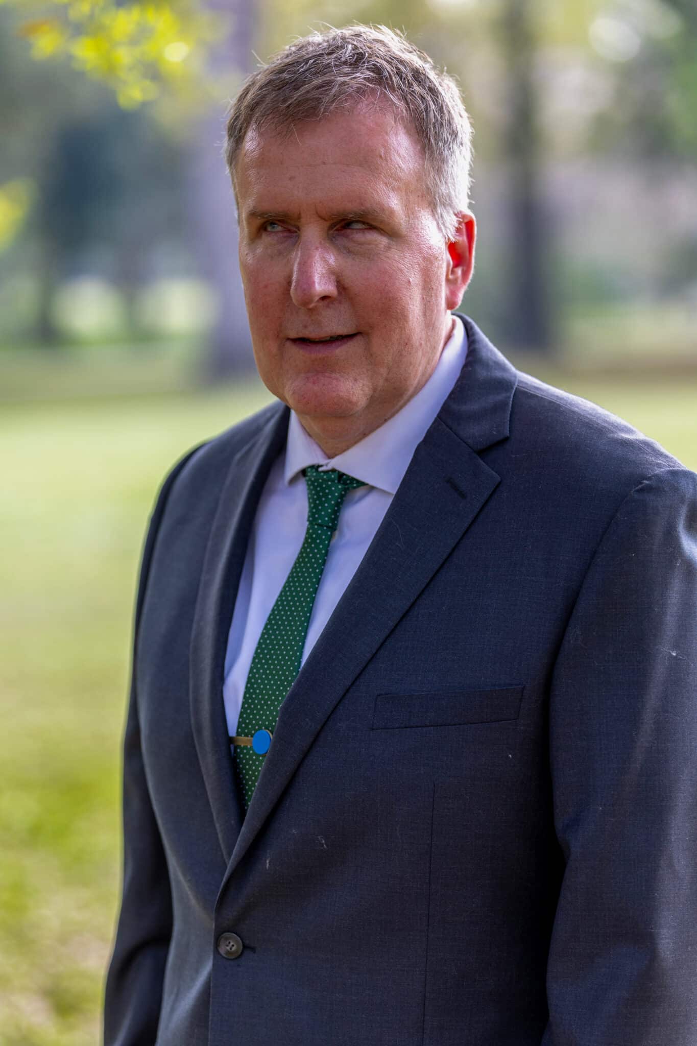 A man in a dark suit with a green tie standing outdoors in a park.