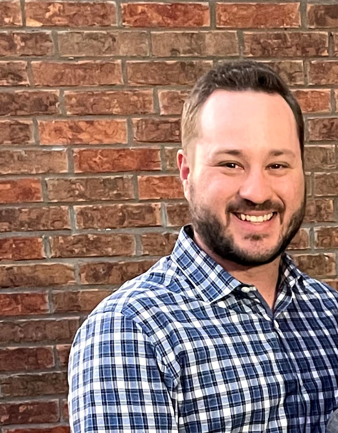 A smiling man with short hair and a beard wearing a blue plaid shirt standing in front of a brick wall.